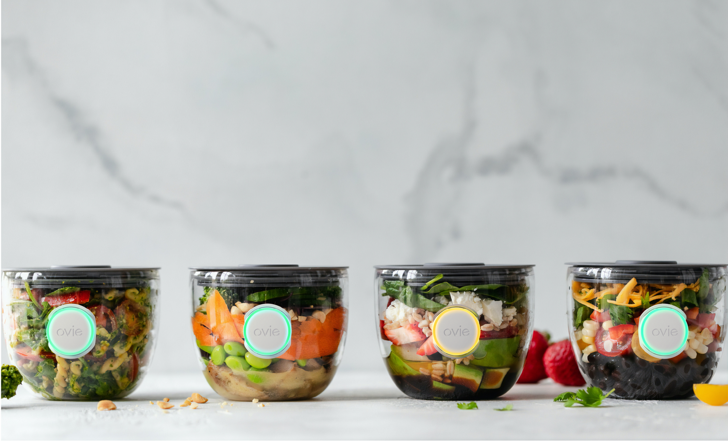 Image of 4 food prep containers lined up on a marble counter with lit up Ovie LightTags on the front of each container. 3 of the LightTags are lit up Teal. One is lit up yellow.