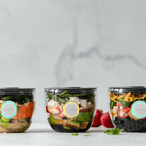 Photo of 3 food prep containers sitting on all marble counter with marble background. Each container has an Ovie LightTag lit up on the face of the container-- the middle LightTag is lit up yellow
