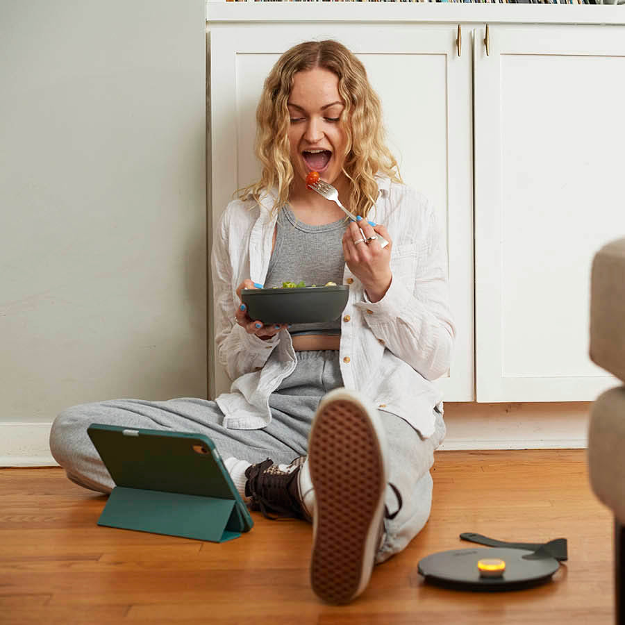 Photo of hip blond Gen Z woman looking at her ipad while sitting on the floor and eating a salad out of a porter bowl. Top of the porter container is sitting on the floor next to her and has an Ovie LightTag on top that is lit up yellow