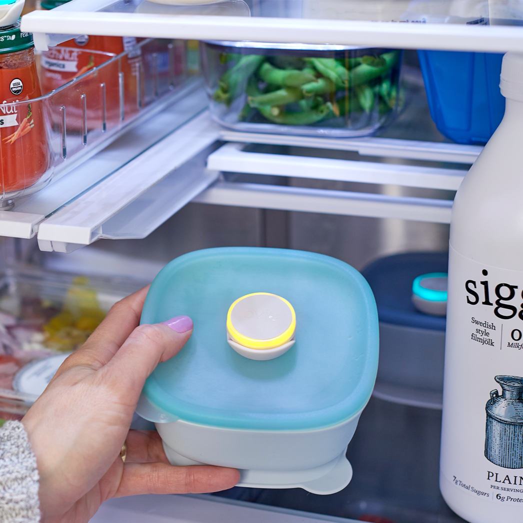 Woman grabbing container with LightTag on top that is lit up yellow. Other LightTags on foods in the fridge are teal.