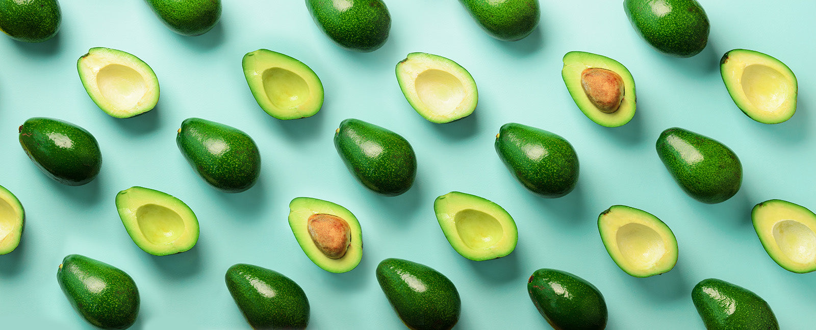 Bright and fun image of avocados that are whole and cut in half laid out in pattern on light teal background