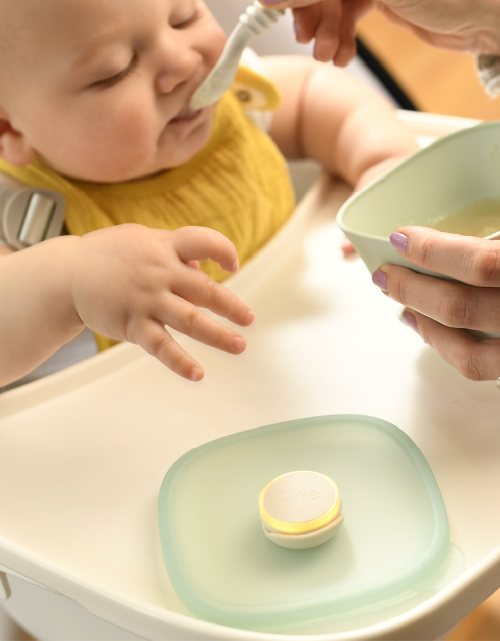 Photo of cute baby in high chair being fed homemade applesauce out of food storage container. Container lid with Ovie LightTag lit up yellow is sitting on high chair tray