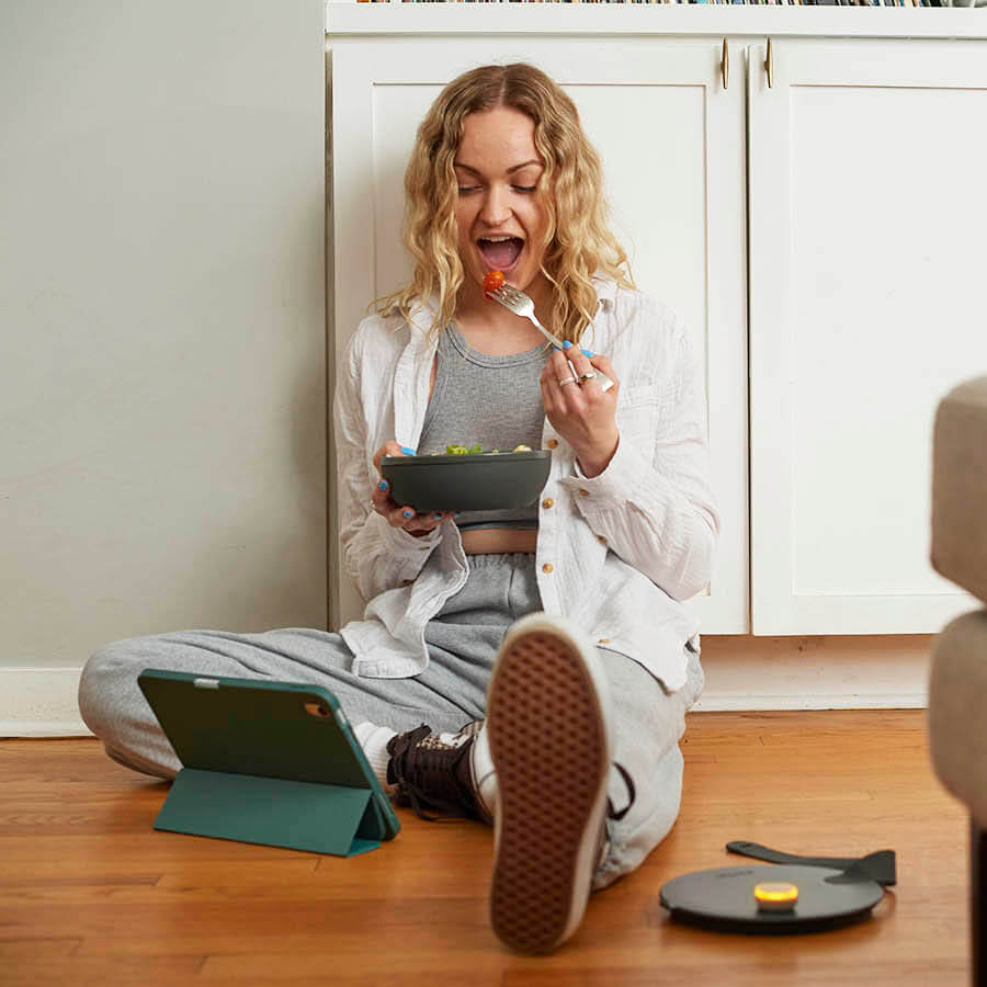 Photo of cute, hip, blond, Gen Z woman happily sitting on the floor, eating salad and watching her ipad. The lid to her food container is sitting next to her on the floor with an Ovie LightTag on top that is lit yellow.