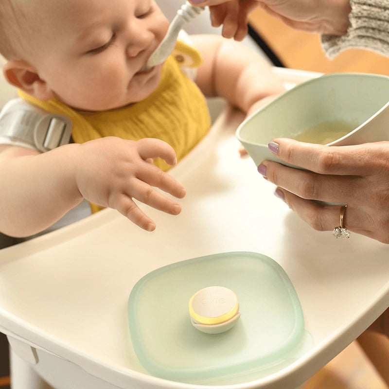 Photo of cute baby in high chair being fed homemade applesauce. Lid to applesauce container is sitting on high chair with Ovie LightTag lit up in yellow on top.