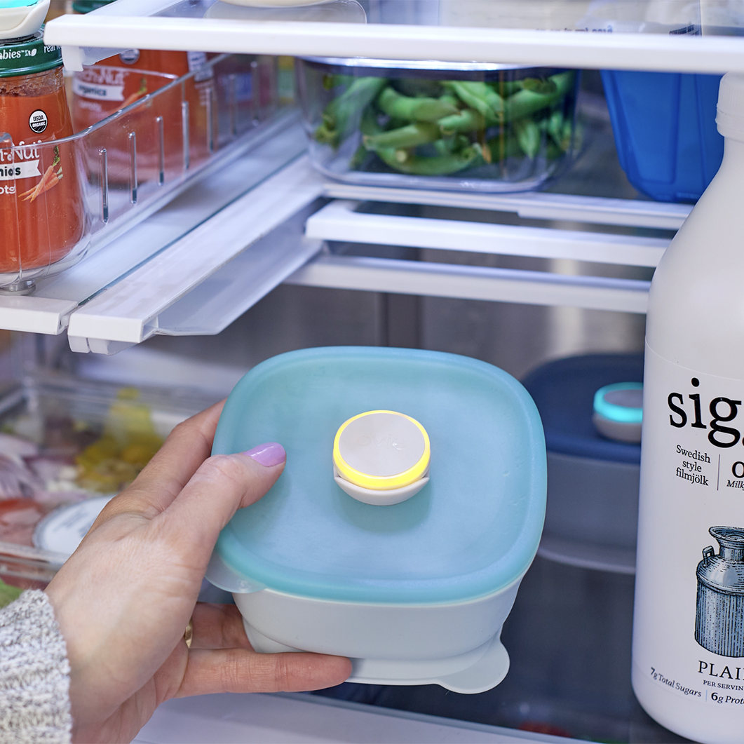 Photo of woman taking small baby food container with Ovie LightTag lit up yellow on it. There are other foods with and without LightTags around fridge.
