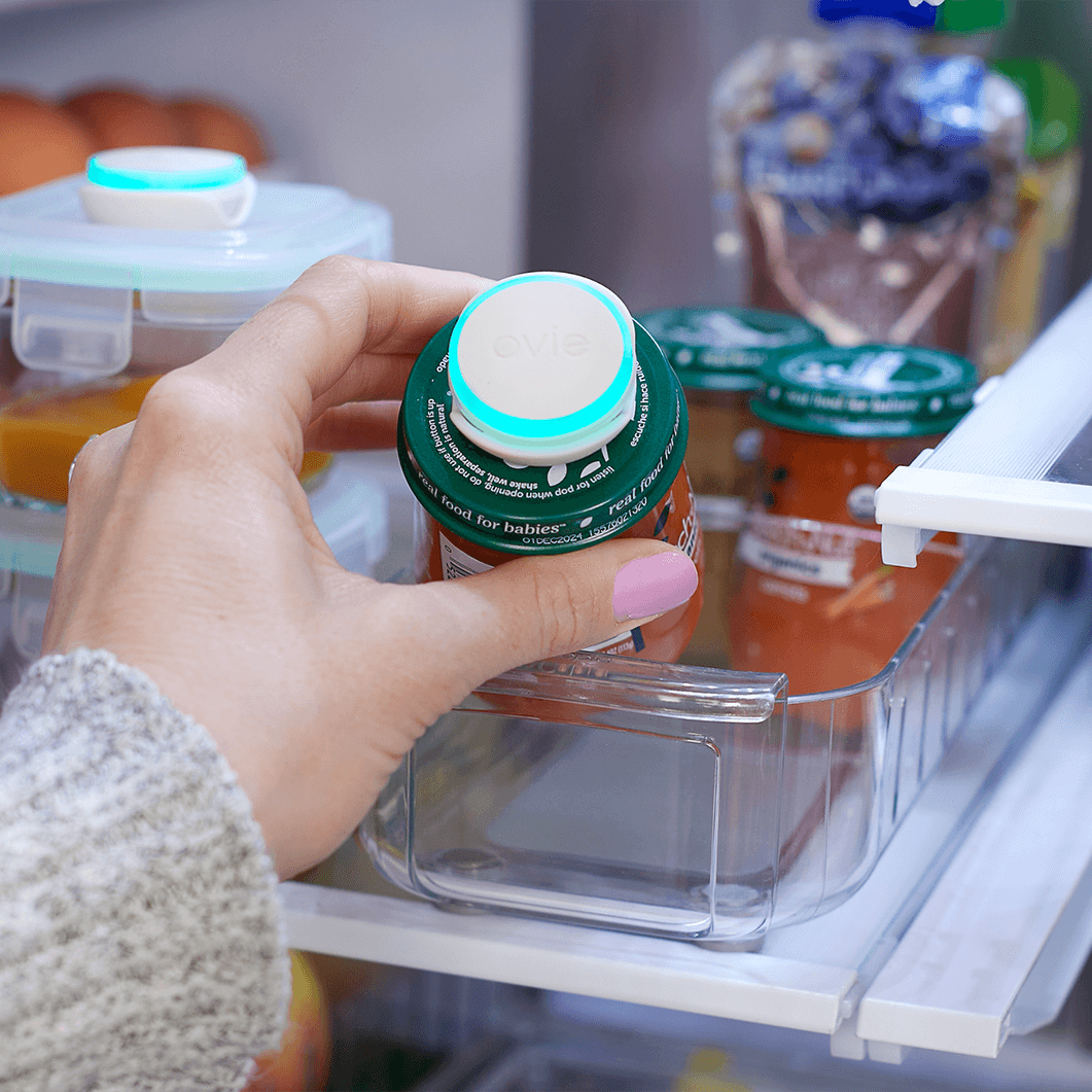 Photo of woman reaching into the fridge and grabbing a jar of baby food with an Ovie LightTag on the lid that is lit up teal.