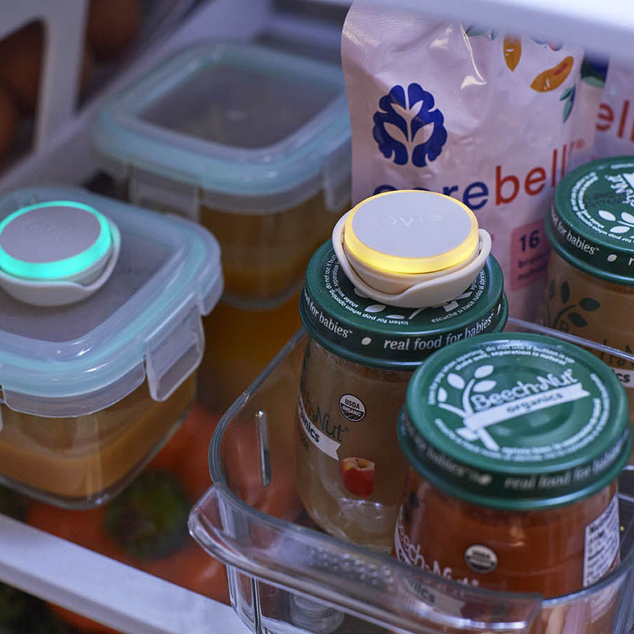 Photo of various baby food containers and jars in fridge. Two containers have Ovie Lightatgs on the top of them. The light on the BeechNut jar is lit yellow.