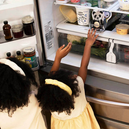 Photo of two cute girls with curly hair in yellow dresses reaching into fridge for animal-shaped food pouches with Ovie LightTags on them
