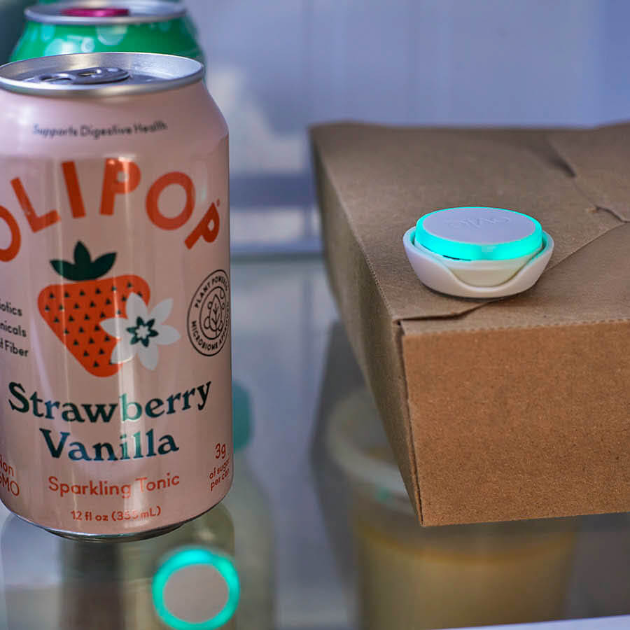Close up photo of a fridge shelf with olipop soda and takeout container. Ovie LightTag lit up teal on top of the takeout container and one LightTag can be seen through the shelf to the shelf below where it is on a dressing container