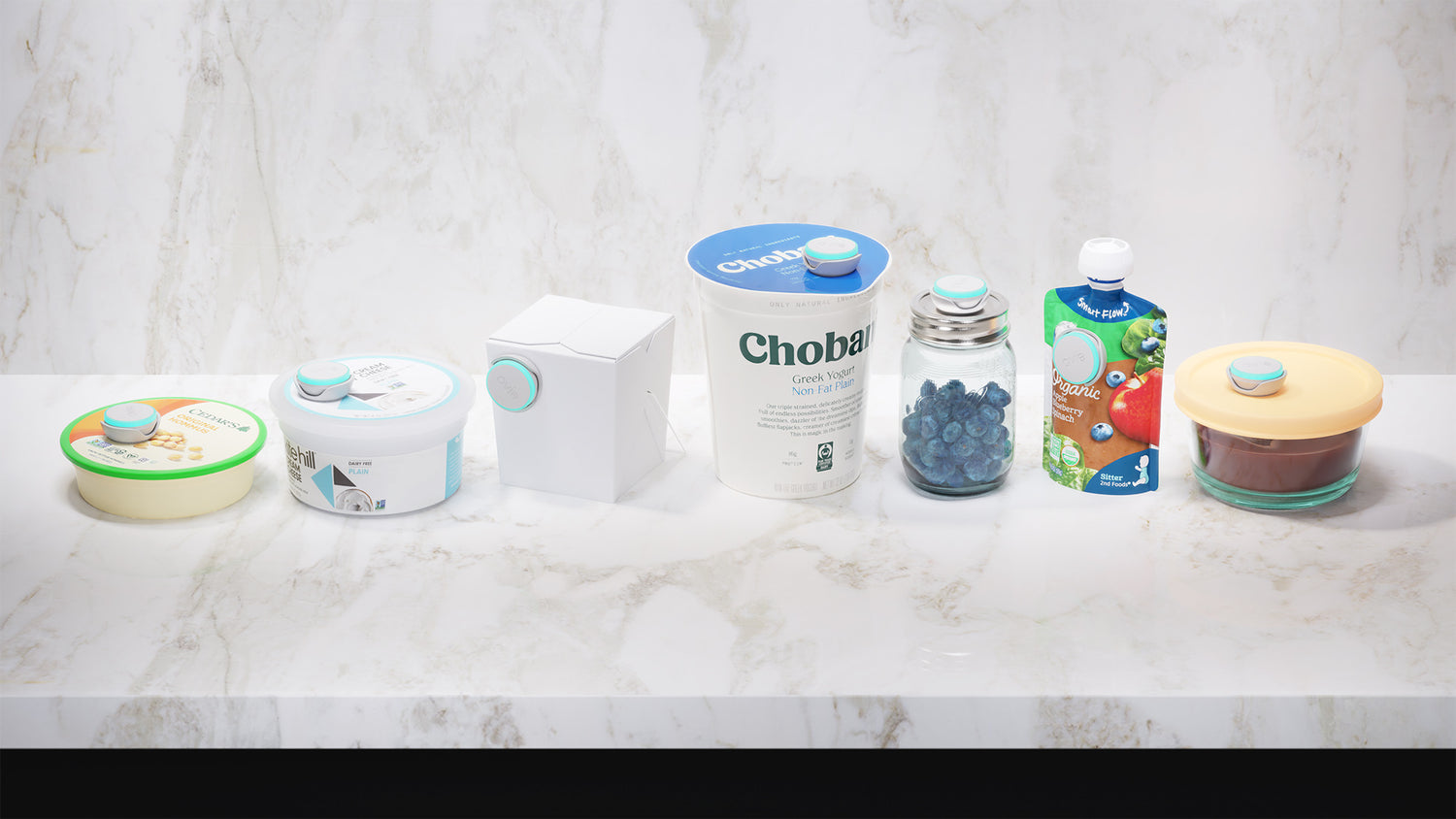Image of 7 different food containers lined up on marble counter with marble backsplash. Each container from hummus to puree has an Ovie LightTag attached to it either on the top or the side