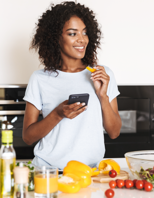 Photo of cute, smiling, dark skinned millennial woman standing in white and black kitchen holding a bit of food in one hand and her phone in the other. 