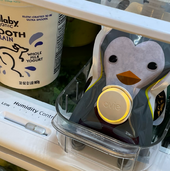 Photo of cute baby food pouch shaped like a penguin the fridge with yellow-lit Ovie LightTag attached to the front 