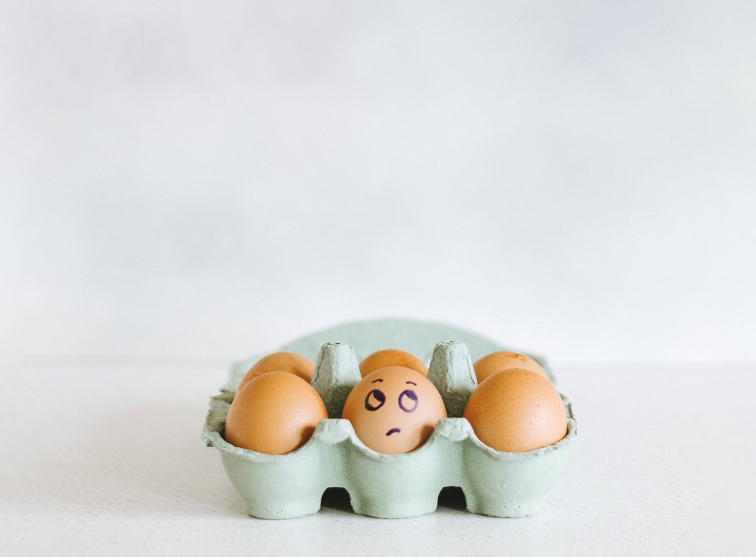 Photo of light aqua-gray 6-egg carton full of brown eggs on an all white background. Middle egg has a skeptical face drawn on the front in sharpie